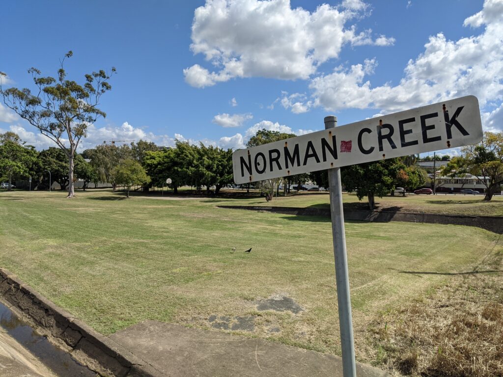 A sign reads "Norman Creek" in front of an empty grassy parkland