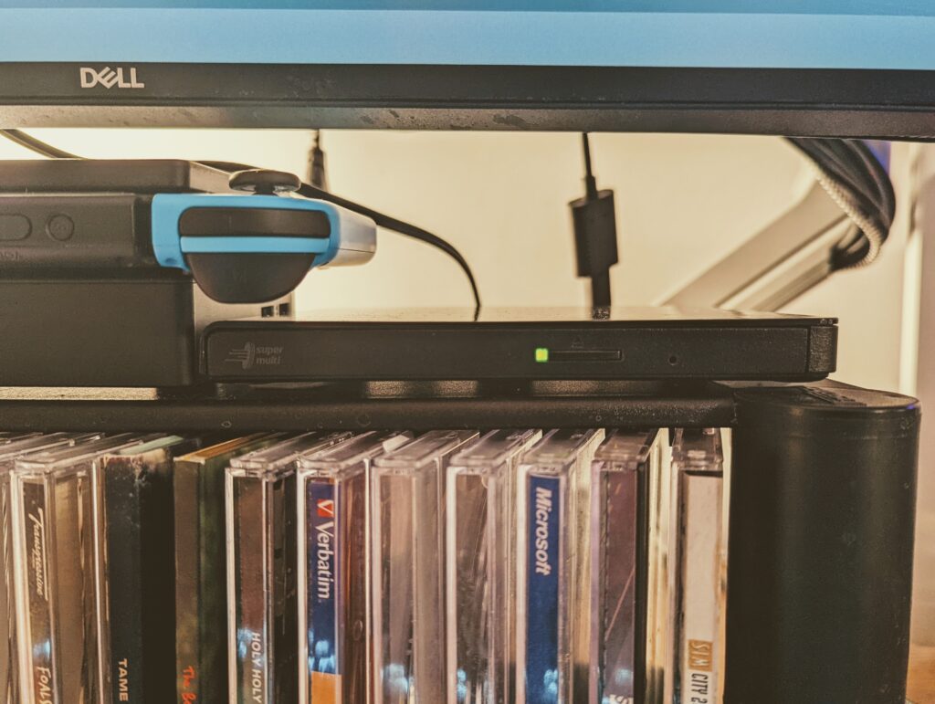 A slimline USB CD drive underneath a monitor and charging Nintendo Switch, with some jewel cases underneath