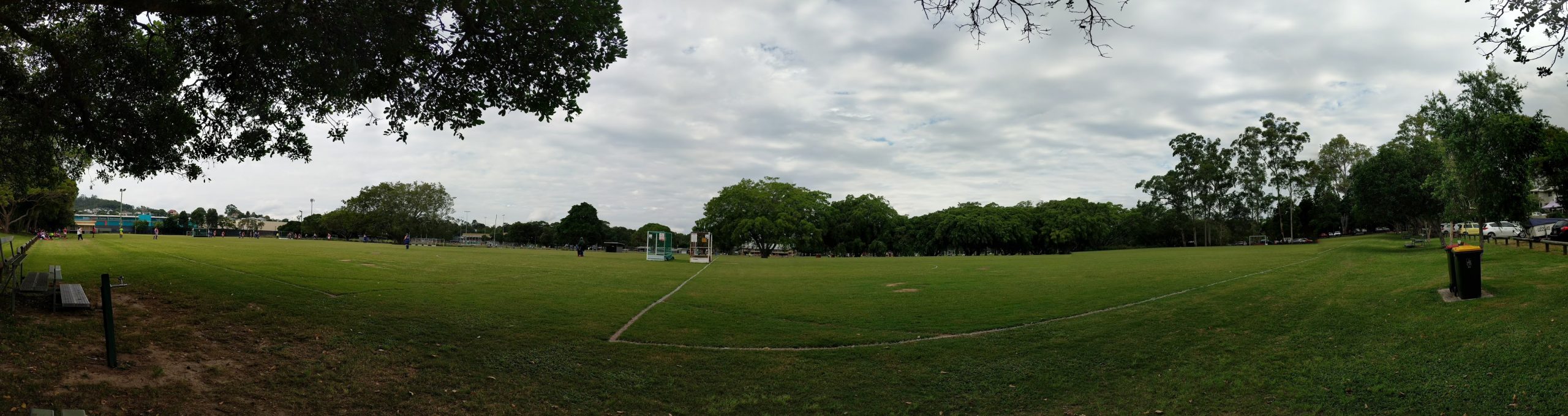 A panorama of a field with trees overhanging. People in uniform playing hockey to the left, and a pair of trash and recyling bins to the right.