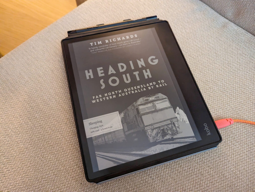 A Kobo ereader on the couch showing Tim Richards Heading South book in black & white. there's a bright pink cable coming out the side.