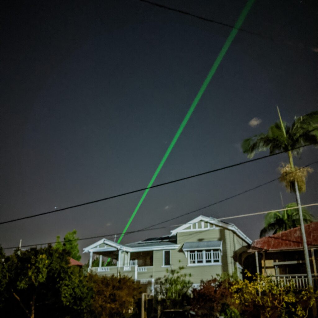 A green laser shoots across the night sky, over old wooden houses.