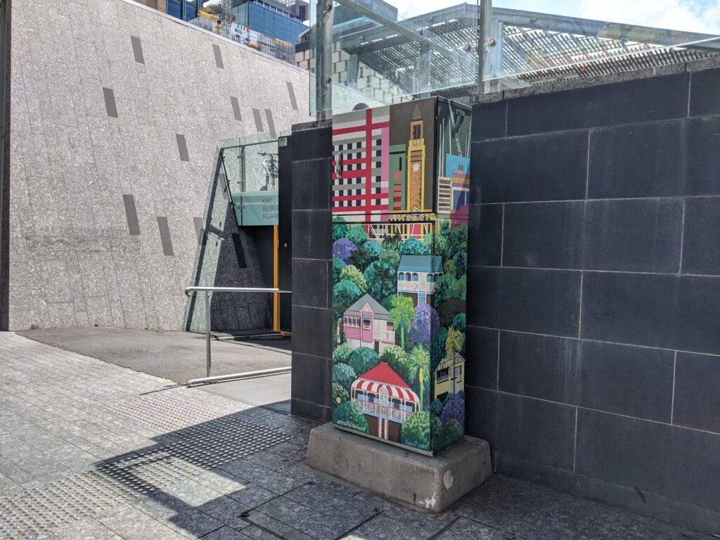 A grey, concrete jungle, with a brightly coloured traffic signal box about as tall as a person. The box is covered with leafy green suburban queenslander houses with high rises in the background.