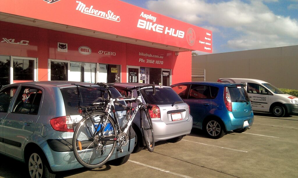 A black & white bike on the back of a blue Hyundai Getz, in front of the red facade of the Aspley Bike Hub (no longer there)
