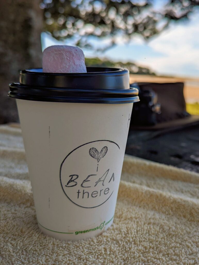 A disposable coffee cup printed with "Bean There" and a love heart. A pink marshmallow on top, and the beach in the distance.