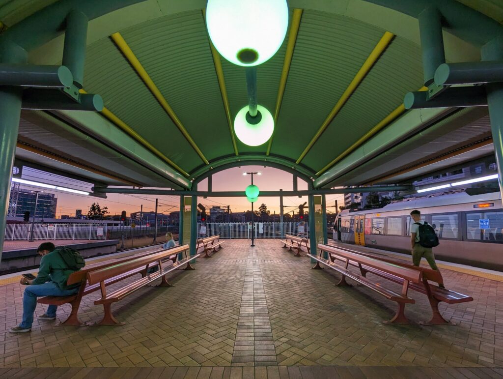 Looking out of the station at Roma Street. the corrugated shelter arches overhead. Greenish flourescent orbshang from the roof, as the sun sets outside.
