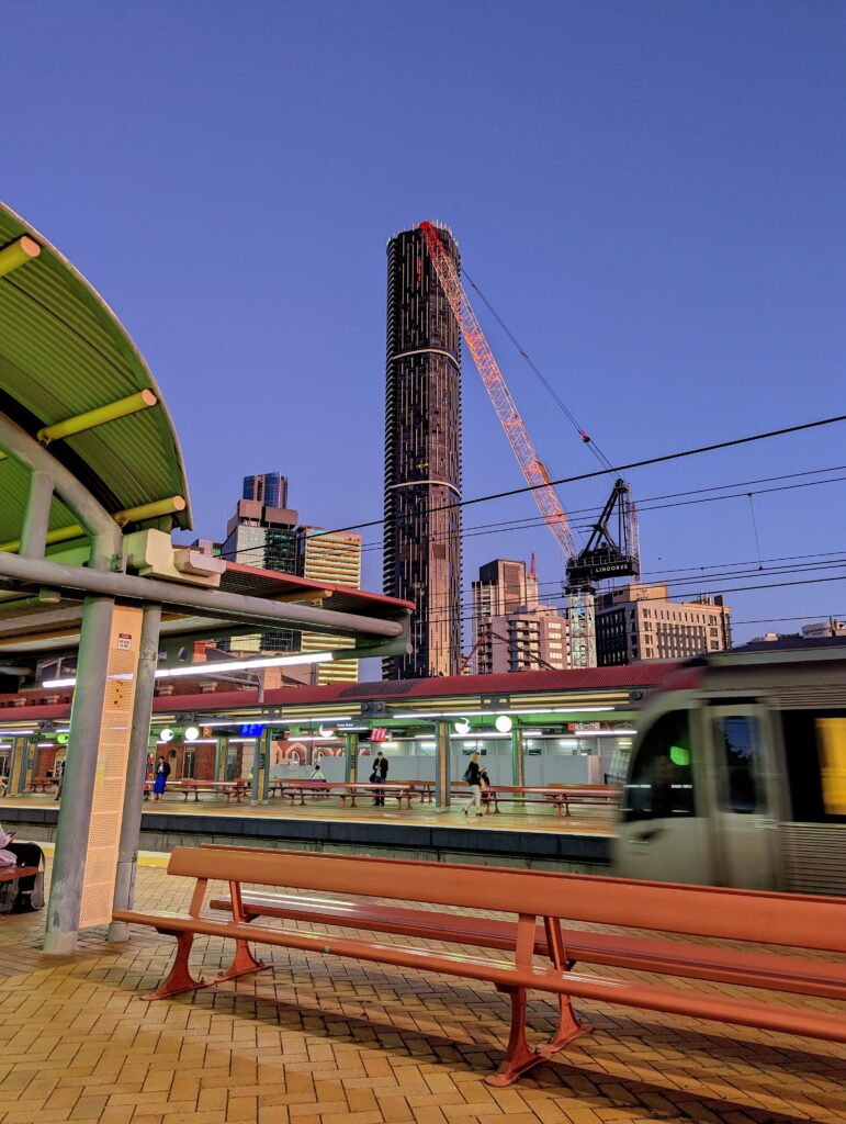 A train rushes past at the platform, the curved corrugated iron is an eerie green colour from the flourescent lights. The Meriton towers in the background.