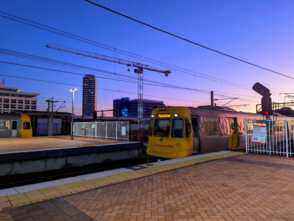 An older Ferny Grove train pulls into the station, as the sun sets in the distance. The sky is mostly purple, but the reflection in silver the train is pink and gold.