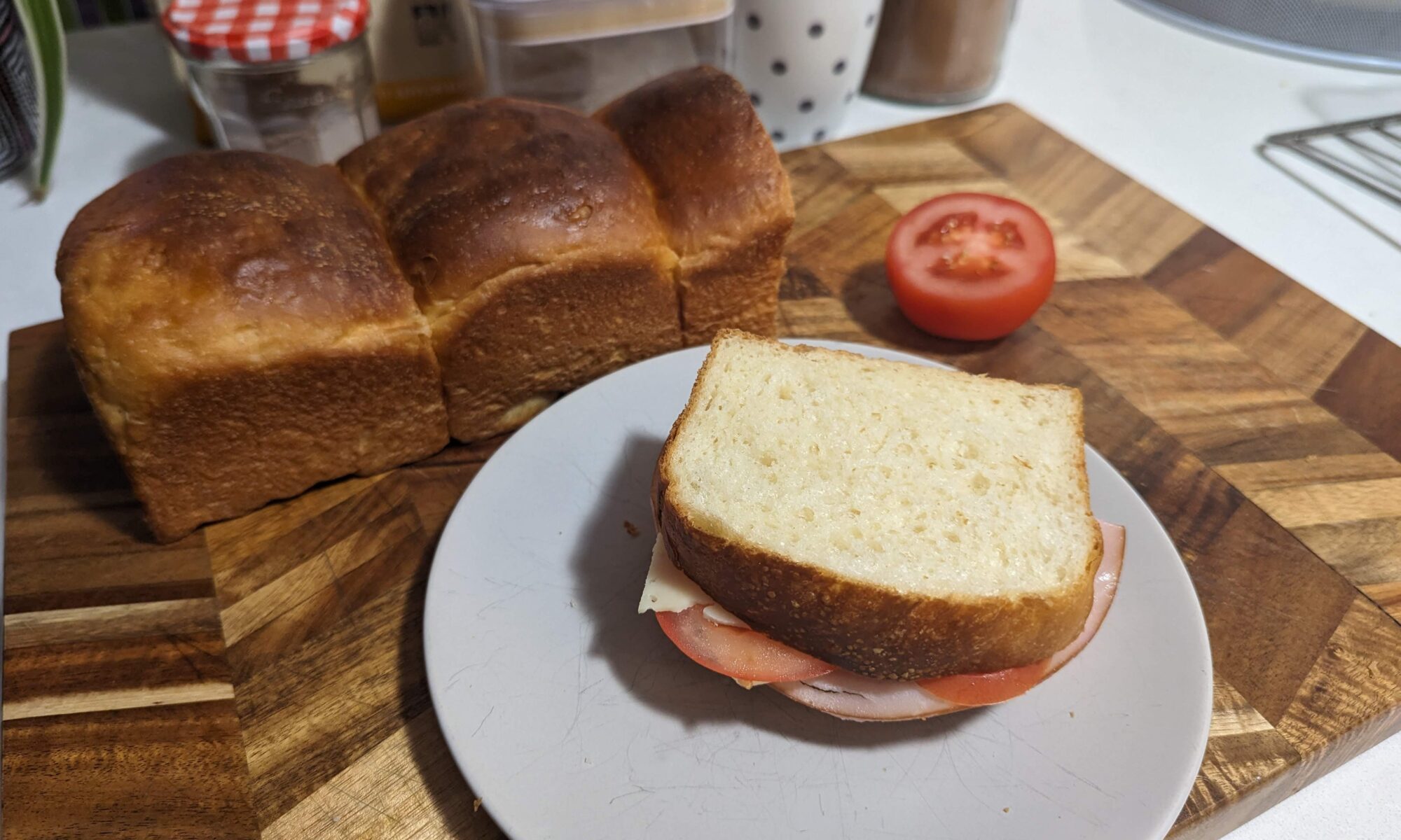 A loaf of delicious golden milk bread, with a freshly made ham cheese and tomato sandwich in the foreground. It looks succulent.