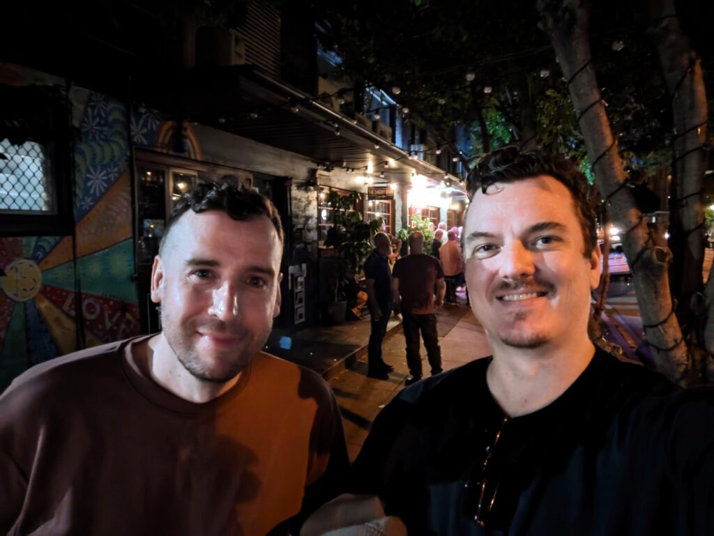 Dan and I, two guys taking a selfie on the street in front of a crowded but not very in-focus bar.