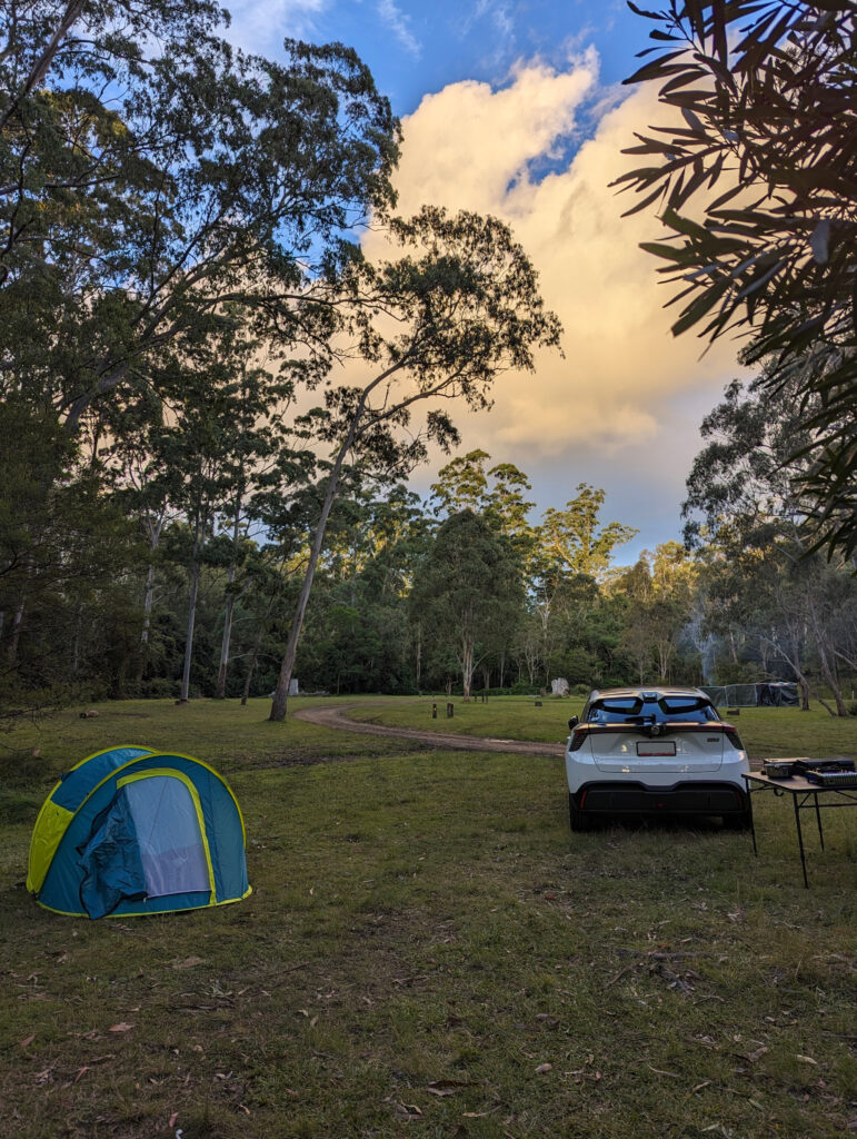 a car and a tent and a little table covered in things in a campground, with lots of trees and a little dirt path