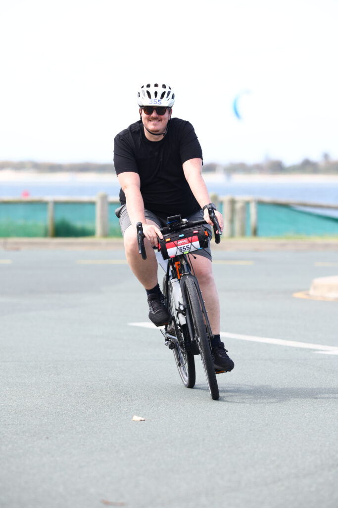 Man on bike, smiling, there's a blurry windsurfer in the background.