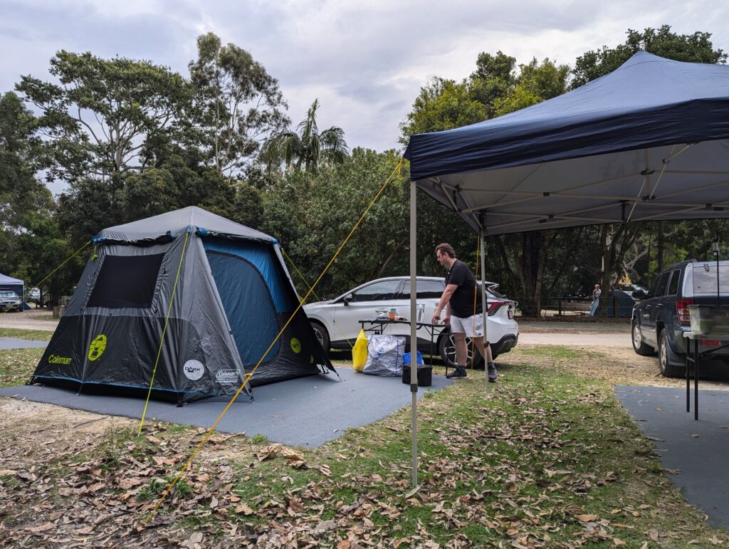 A square pyramidal looking tent and gazebo pitched on a grassy space with autumnal leaves everywhere. A man (meeee) lifts one edge of a camp table.