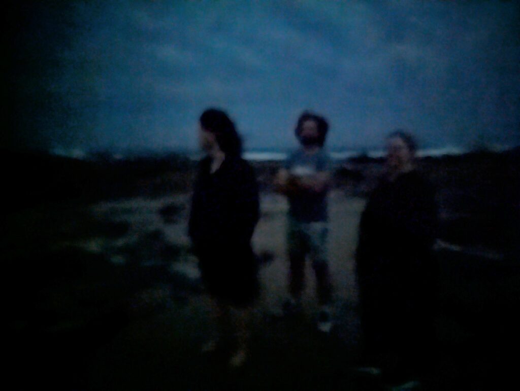 It's blurry and dark. Three ghostly figures stand on a barely discernible beach at night. One woman looks aloof away from the camera, a man has his arms crossed.