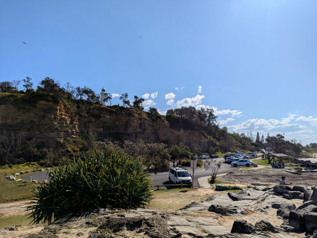 A lighthouse sits atop a cliff. A car park sits at the foot with a bunch of cars, trees and rocks scattered about.