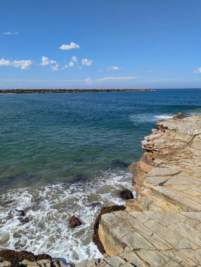 Looking out over a cliff to the ocean below. The water goes from blue to green to sandy and washy as it crashes into the rocks.