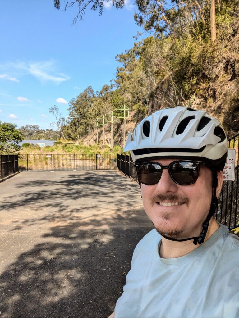 Man with helmet and scratched sunglasses stands in front of a cliff and an overgrown wharf that you can't really tell what it is.