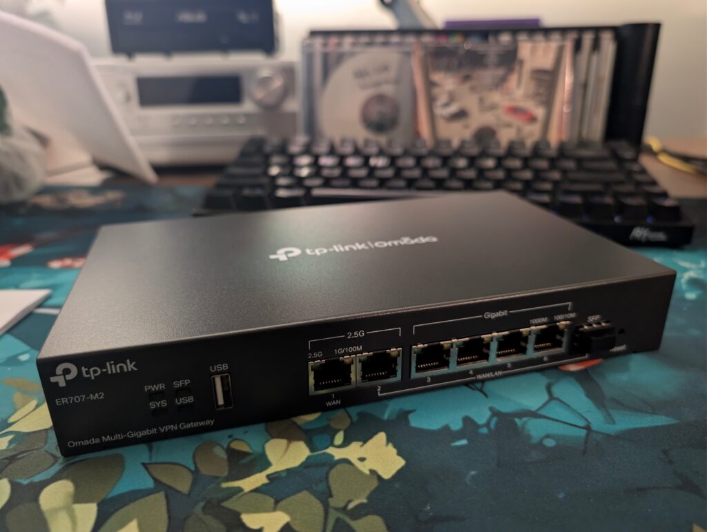 The TP-Link Omada ER707-M2 sitting on a desk. It has two 2.5gbit ports, 5x 1 gbit ports, and a USB connection on the front. It's just a sleek black metal box, like an old Netgear router.